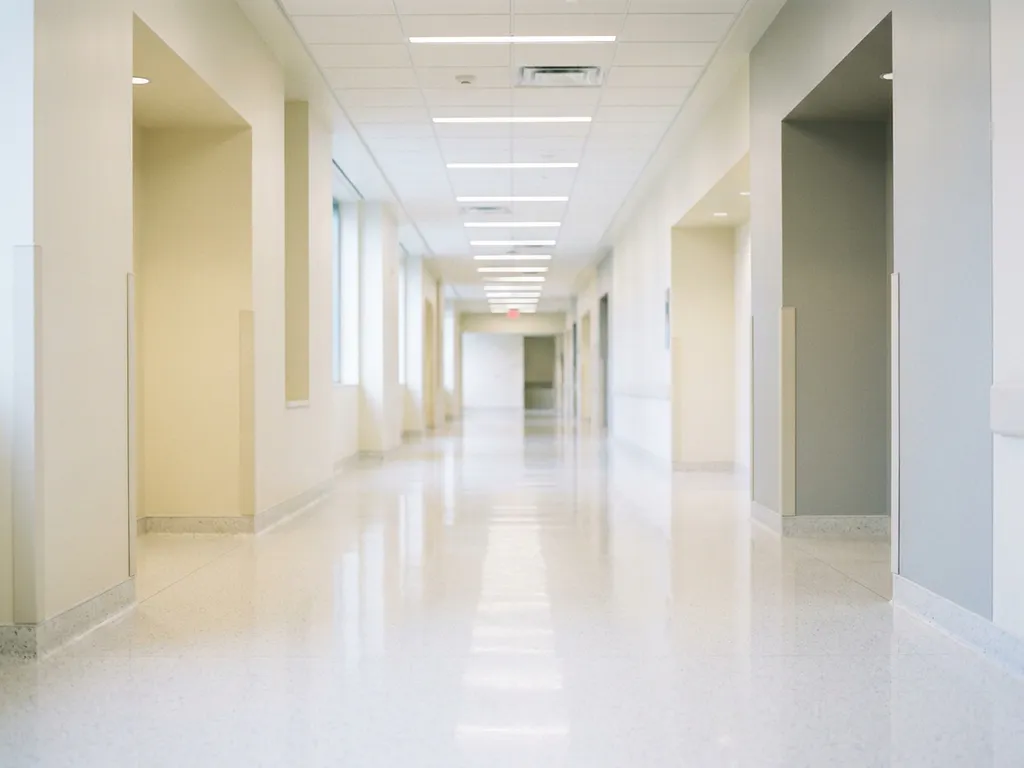 Leerer Krankenhausflur mit glänzendem weißem Boden, neutralen Wänden und hellem Deckenlicht, das in die Tiefe führt.
