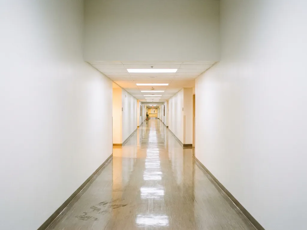 Langer Krankenhausflur mit poliertem Linoleumboden, der Neonlicht reflektiert, leichte Schmutzspuren im Vordergrund sichtbar.
