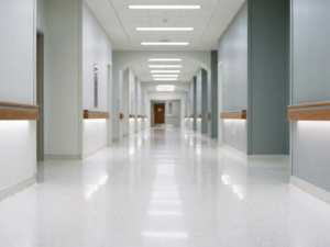 Leerer Krankenhausflur mit glänzendem weißem Boden und Neonbeleuchtung, die sich in kühlen Grau- und Weißtönen in die Tiefe erstreckt.