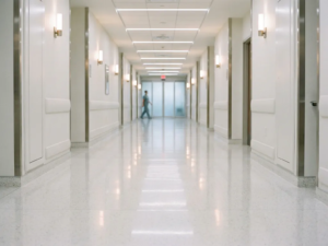 Weitläufiger Krankenhausflur mit glänzendem weißen Boden, der Neonlicht reflektiert, und modernen klinischen Wänden in kühlen Grautönen.