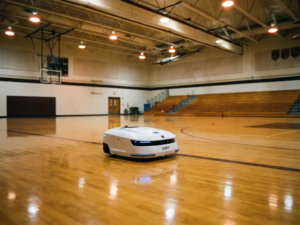Weißer Reinigungsroboter fährt autonom über den polierten Boden einer leeren Turnhalle mit Basketballmarkierungen.