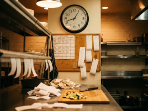 Unübersichtliche Restaurantküche mit Bestellzetteln, handgeschriebenem Dienstplan und Wanduhr über halbgeschnittenem Gemüse auf Holzbrett.